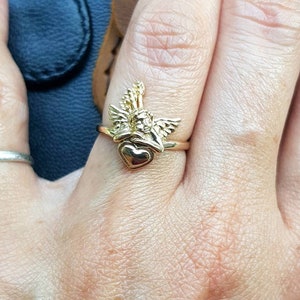 May include: A gold-tone ring featuring a cherub holding a heart. The ring is worn on a person's finger.