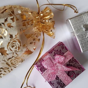 May include: Three gift boxes, one in a gold and white floral pattern, one in a silver and white patterned design, and one in a pink and white floral pattern with a pink ribbon.