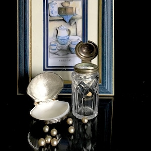 Op de afbeelding: Een stilleven met een ingelijste prent van blauw en wit keukengerei, een heldere glazen pot met een metalen deksel, een zilveren schelpvormige container en verschillende parels. De pot heeft een diamantgeslepen patroon. De prent heeft de tekst "SWR1-145".