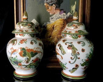 May include: Two white porcelain ginger jars with lids, decorated with green, orange, and red floral and bird designs. The jars are sitting in front of a framed needlepoint picture.