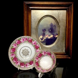 May include: A vintage teacup and saucer with a pink rose pattern. The saucer has a gold rim and a gold design in the center. The teacup has a pink rose pattern and a gold rim. The teacup and saucer are on a black surface in front of a framed painting of a still life.