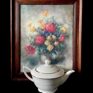 May include: A white porcelain teapot with gold trim and a decorative lid. The teapot is in front of a framed floral painting with red, yellow, and blue flowers. The frame is made of dark wood.