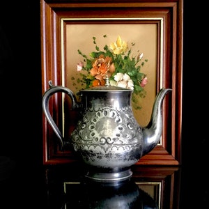 以下が含まれることがあります： 装飾的なデザインが施された銀製のティーポットと、背景に花の絵画