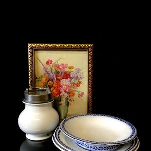 May include: A white ceramic jar with a silver lid and a blue and white patterned plate and bowl. The jar is sitting on a black surface in front of a framed picture of a bouquet of flowers.
