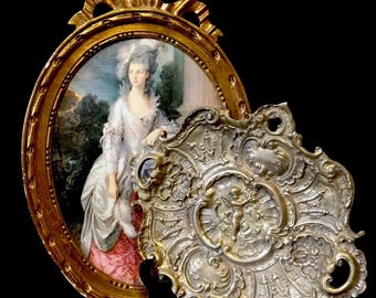 May include: An antique gold-framed portrait of a woman in a white dress, next to an ornate silver-colored tray. The portrait has an oval shape with a bow at the top. The tray features a cherub design.