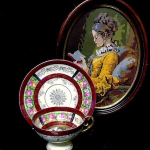 May include: A vintage tea set with a cup, saucer, and plate, featuring a burgundy rim and floral design. A framed needlepoint portrait of a woman reading is in the background. The cup has a gold handle.