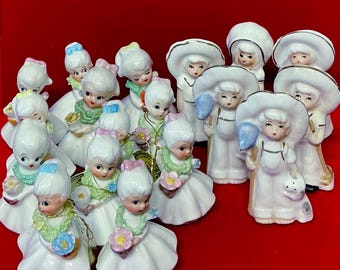 May include: A collection of vintage porcelain figurines of young girls in white dresses and bonnets. Some hold flowers or balloons, others have wide-brimmed hats. The figurines have painted facial features and are arranged on a red surface.