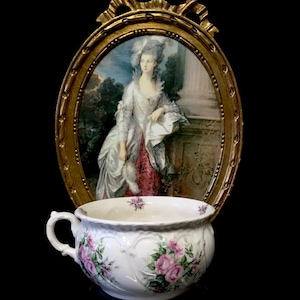 Może przedstawiać: Biały nocnik z porcelany z kwiatowym wzorem z różowymi różami. Nocnik ma jedną rączkę i jest ozdobiony wypukłym, ozdobnym wzorem. Nocnik stoi przed złotą owalną ramą z obrazem kobiety w białej sukni.