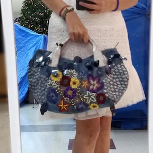 May include: A woman in a white dress holds a denim handbag with colourful embroidered flowers and sequins. The bag has a silver handle and a white lining.