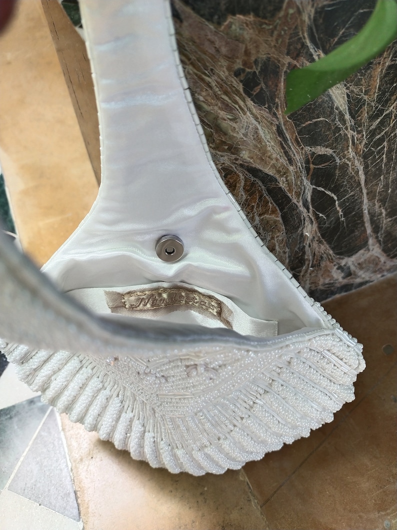 May include: A white beaded evening bag with a satin lining and a metal clasp. The bag is shaped like a seashell and has a long strap.