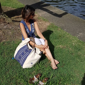 May include: A woman sits on the grass wearing a blue and white patterned top and white shorts. She is holding a large white tote bag with blue and white patterns. The bag is sitting on the grass next to her. She is wearing sandals.