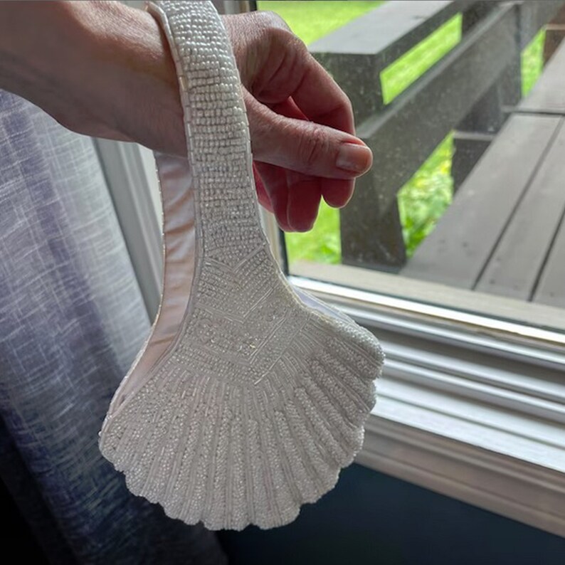 May include: A white beaded handbag shaped like a seashell. The bag has a beaded handle and a white satin lining.