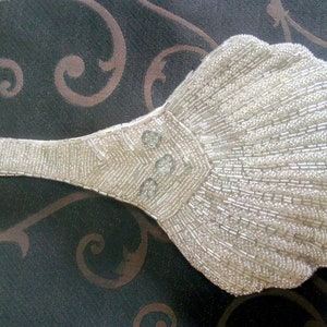May include: A white beaded clutch shaped like a seashell with a long handle. The clutch has a pearl-like sheen and is decorated with three round, clear stones.