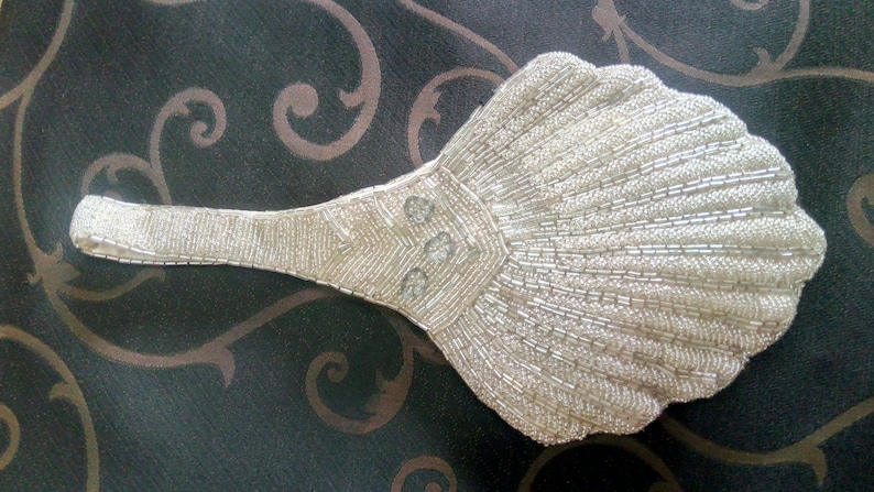 May include: A white beaded clutch shaped like a seashell with a long handle. The clutch has a pearl-like sheen and is decorated with three round, clear stones.