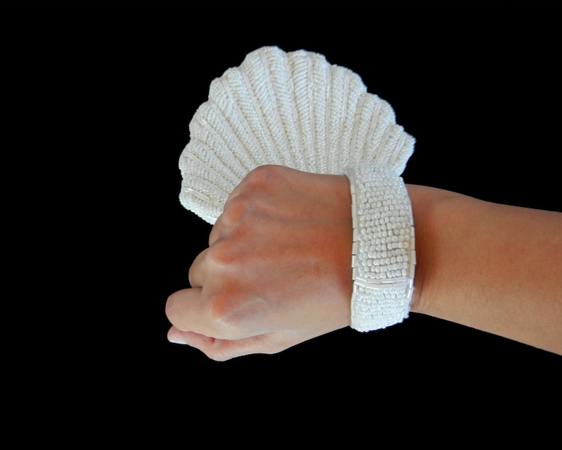 May include: A white beaded bracelet with a shell-shaped design. The bracelet is made of small, white beads that are strung together to create a delicate and elegant look.