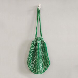K&ouml;nnte beinhalten: Eine gr&uuml;ne Perlen-Kordeltasche mit silbernen Akzenten. Die Tasche h&auml;ngt an einem Haken an einer wei&szlig;en Wand.