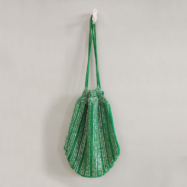 K&ouml;nnte beinhalten: Eine gr&uuml;ne Perlen-Kordeltasche mit silbernen Akzenten. Die Tasche h&auml;ngt an einem Haken an einer wei&szlig;en Wand.