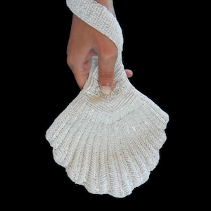 May include: White beaded shell-shaped wristlet purse. The wristlet is made of white beads and has a shell-like shape. It is held in a hand.