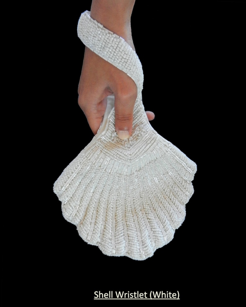 May include: White beaded shell-shaped wristlet purse. The wristlet is made of white beads and has a shell-like shape. It is held in a hand.