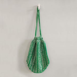 Puede incluir: Una bolsa de cord&oacute;n de cuentas verdes con detalles plateados. La bolsa est&aacute; colgada de un gancho en una pared blanca.