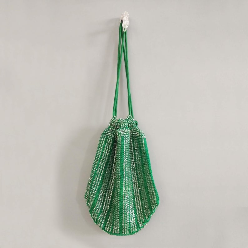 Puede incluir: Una bolsa de cord&oacute;n de cuentas verdes con detalles plateados. La bolsa est&aacute; colgada de un gancho en una pared blanca.