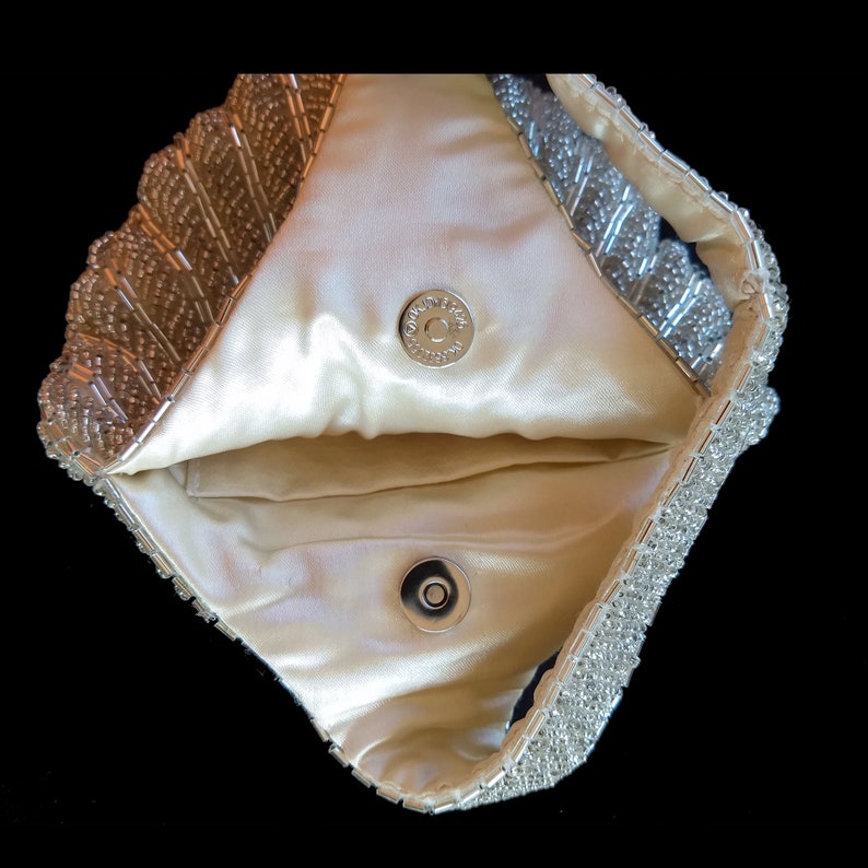 May include: A silver beaded clutch purse with a satin lining and a snap closure. The purse is open and the lining is visible.