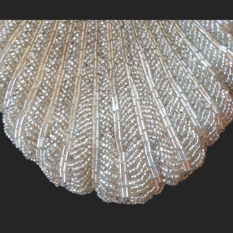 May include: A white beaded clutch purse shaped like a seashell. The purse is made of small, round beads that are sewn together to create a textured surface.
