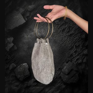K&ouml;nnte beinhalten: Eine silberne, mit Pailletten besetzte Kordeltasche mit zwei goldenen Metallringen als Henkel.