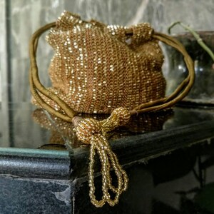 K&ouml;nnte beinhalten: Eine goldene Perlen-Kordeltasche mit Quaste. Die Tasche besteht aus kleinen, gl&auml;nzenden Perlen und hat einen langen, d&uuml;nnen Riemen.