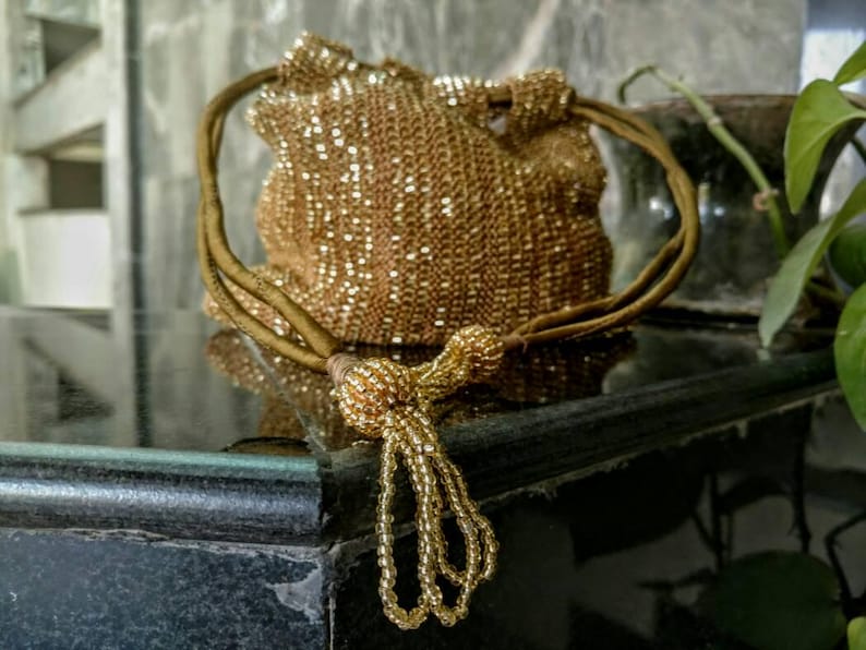 K&ouml;nnte beinhalten: Eine goldene Perlen-Kordeltasche mit Quaste. Die Tasche besteht aus kleinen, gl&auml;nzenden Perlen und hat einen langen, d&uuml;nnen Riemen.