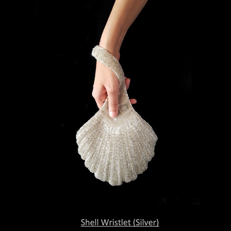 May include: A silver beaded wristlet shaped like a seashell. The wristlet has a beaded strap and is held by a hand.