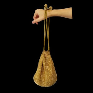 Puede incluir: Una bolsa de cord&oacute;n de cuentas doradas con una correa larga y delgada. La bolsa cuelga de la correa, que est&aacute; sostenida por una mano.