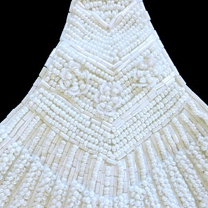 May include: Close-up of a white beaded garment with a chevron pattern and floral embellishments.