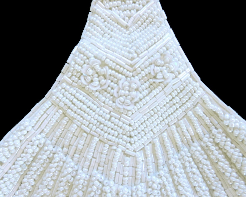 May include: Close-up of a white beaded garment with a chevron pattern and floral embellishments.