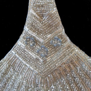 May include: A close-up of a silver beaded embellishment with a geometric design. The embellishment is made of small, round beads and has a pointed shape.