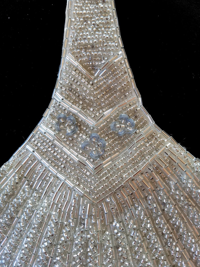 May include: A close-up of a silver beaded embellishment with a geometric design. The embellishment is made of small, round beads and has a pointed shape.