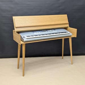 Op de afbeelding: Een houten keyboardstandaard met een neerklappend deksel. De standaard is gemaakt van licht hout en heeft een zilveren keyboard erop.