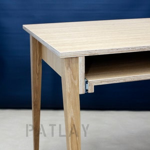 Op de afbeelding: Een licht houten bureau met een uittrekbare lade en vier poten. Het bureau staat voor een blauwe muur met het woord "PATLAY" erop gedrukt.