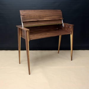 Può includere: Una scrivania in legno con un piano ribaltabile, realizzata in legno di noce con una bordura in legno chiaro. La scrivania ha quattro gambe e si trova su una superficie marrone chiaro.