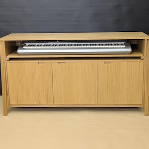 May include: A light oak wood console cabinet featuring a sliding keyboard tray shelf positioned below the top surface. The cabinet includes three vertical cabinet doors with recessed handles, housing a digital keyboard instrument on the tray.