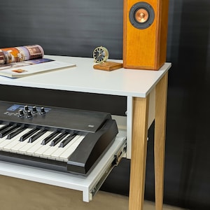 May include: A white desk with light wood legs, featuring a pull-out keyboard tray. A black keyboard is partially extended. A wooden speaker, a small clock, and a magazine are on the desk. The background is black.