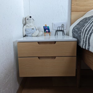 May include: A wall-mounted floating nightstand with two light wood drawers featuring integrated horizontal handles and a smooth white top surface. On the surface sits a plush white dog toy, a small canvas on a wooden easel, and a black wire frame.