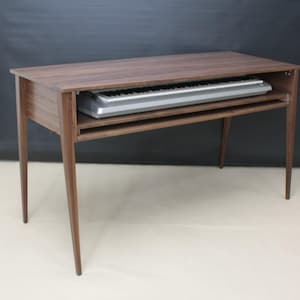 May include: A dark brown wooden keyboard stand with a built-in shelf. The stand has four tapered legs and a rectangular top. A silver and black keyboard is placed on the shelf. The stand is designed to hold a keyboard.