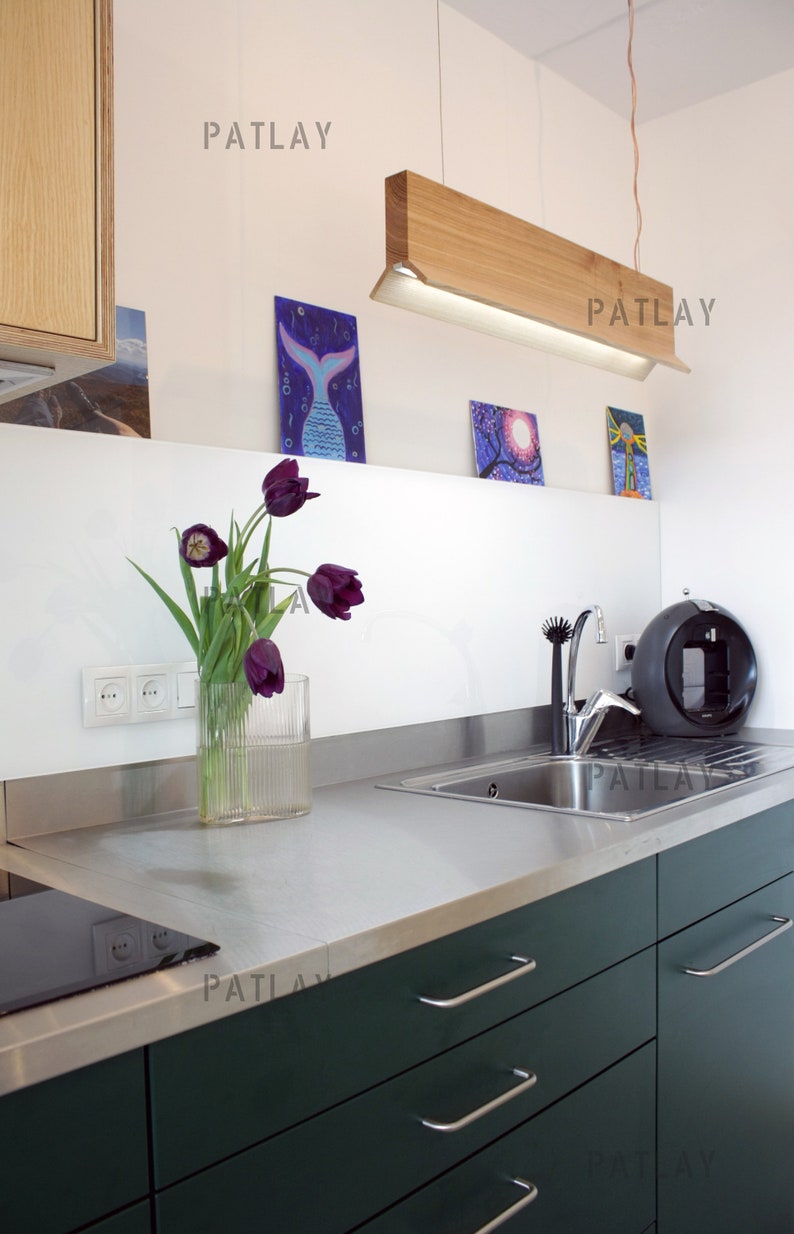 May include: A modern kitchen with dark green cabinets and stainless steel countertops. A wooden light fixture hangs above the sink. There are three framed pictures on the wall behind the sink. A vase of purple tulips sits on the counter.