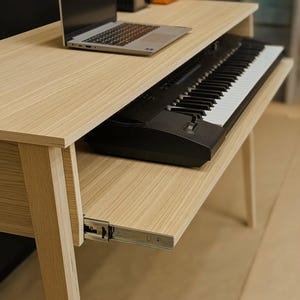 以下が含まれることがあります： 引き出し式のキーボードトレイが付いた明るい木製のデスク。 白いキーが付いた黒いキーボードがトレイにあります。 ラップトップがデスクの上面にあります。 ミニマリストデザイン。