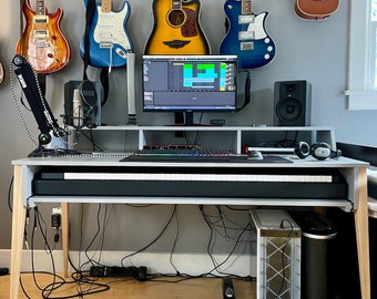 May include: A white desk with a keyboard and a computer monitor. There are five guitars hanging on the wall behind the desk, including an acoustic guitar, an electric guitar, and a ukulele. The desk is in a room with a wooden floor.