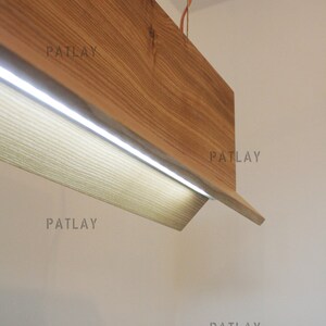 May include: A wooden light fixture with a rectangular shape and a built-in LED light strip. The fixture is suspended from the ceiling by a copper wire. The text "PATLAY" appears five times on the white wall behind the fixture.