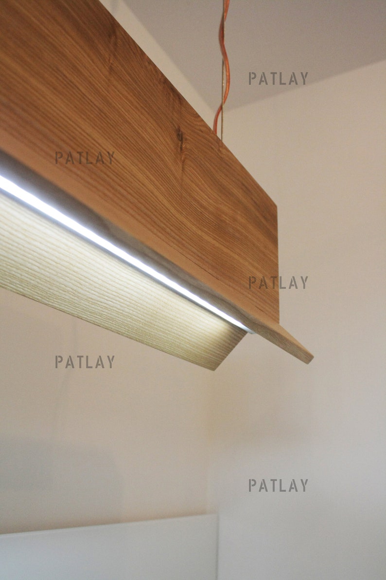 May include: A wooden light fixture with a rectangular shape and a built-in LED light strip. The fixture is suspended from the ceiling by a copper wire. The text "PATLAY" appears five times on the white wall behind the fixture.