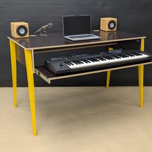 May include: A contemporary music studio desk with a pull-out keyboard tray. The desk has a dark brown top and bright yellow legs. A laptop, speakers, and a small dinosaur figurine are on the desk. A black keyboard is on the pull-out tray.