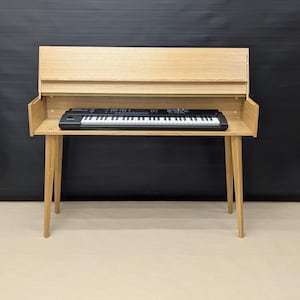 Op de afbeelding: Een licht houten keyboardstandaard met een ingebouwde plank en vier schuine poten. Een zwart-wit keyboard staat op de plank. De standaard heeft een eenvoudig, modern ontwerp.
